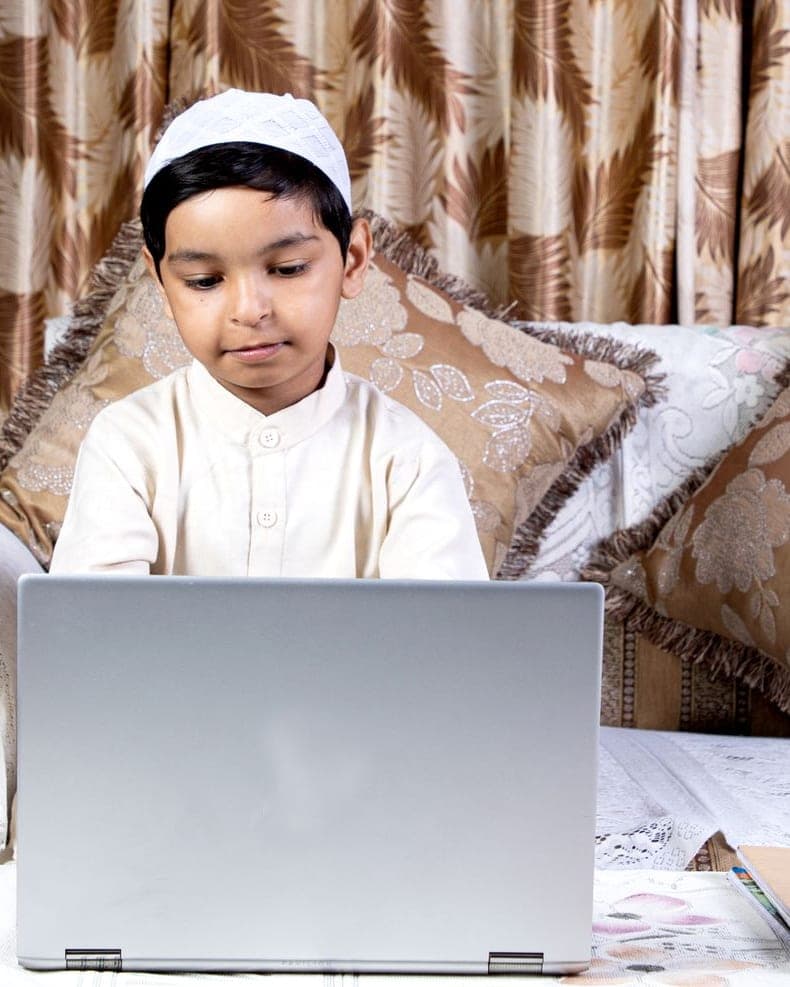 A young boy in a white cap taking an online lesson on a laptop at home